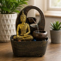 Tabletop Water Feature - 21.5cm - Golden Buddha & Pots showing a golden Buddha statue seated beside cascading water flowing from a small pot into another on a textured base, with plants in the background.