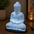 Meditation Buddha Lamp Sky Blue - Oval Stand glowing softly with a crackled texture, placed on a wooden surface with warm ambient lighting and decorative plants in the background.