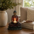 Antique Buddha - Devotee Candle Holder showcasing a serene seated Buddha statue holding a lit candle, placed on a wooden table near books and a textured vase with greenery.