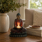 Antique Buddha - Devotee Candle Holder showcasing a serene seated Buddha statue holding a lit candle, placed on a wooden table near books and a textured vase with greenery.