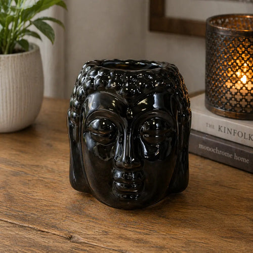 Glossy black ceramic Buddha Oil Burner - Black with detailed facial features, placed on a wooden surface next to a candle holder and potted plant for serene home decor.
