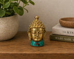 Brass Buddha Figure - Med Head - 8 cm with intricate curls and peaceful expression, adorned with turquoise and red stone details, placed on a wooden surface near books and a plant.