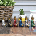 Six Gold Mini Buddha (Assorted Colours) statues in various colors including silver, black, red, blue, and green, displayed on a wooden surface with plants and decorative items in the background.