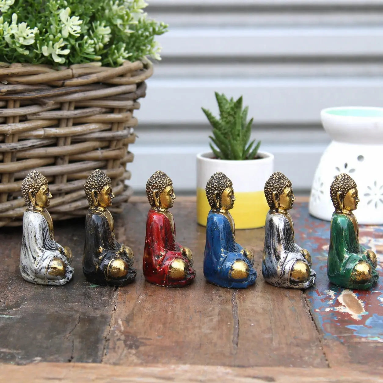Gold Mini Buddha (Assorted Colours) statues in various hand-painted finishes with gold detailing, arranged on a wooden surface with plants in the background as Buddha decor.