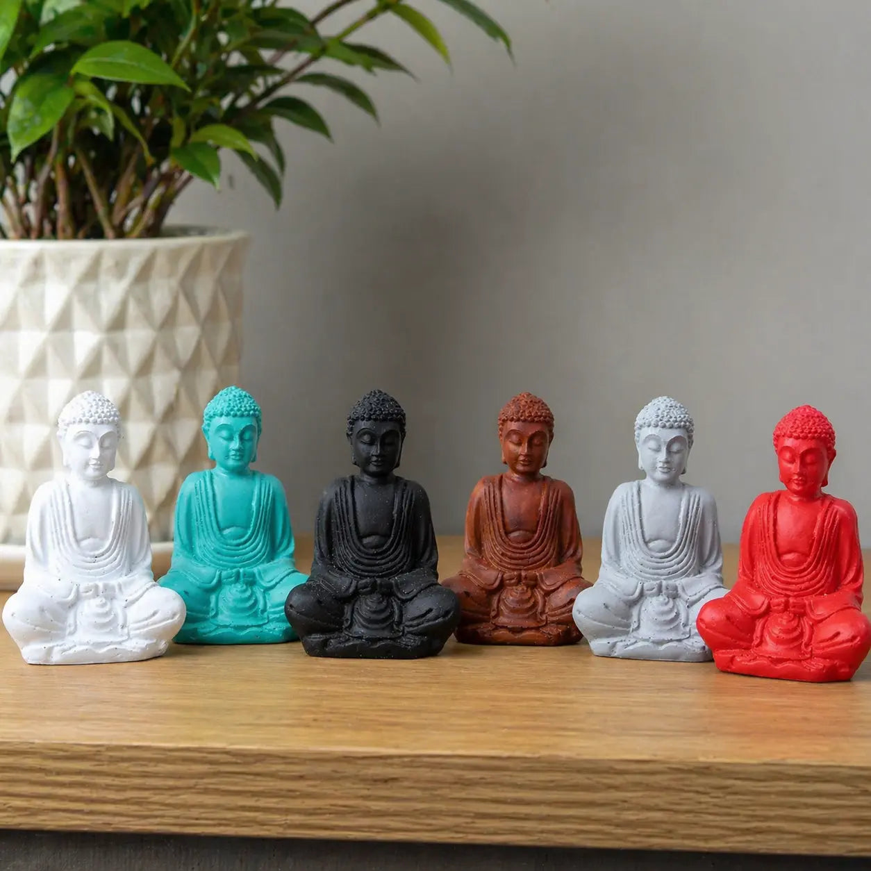 A collection of Matt Mini Buddha (Assorted Colours) statues in white, teal, black, brown, grey, and red, displayed on a wooden surface with a plant in the background.