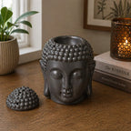 Buddah Head Oil Burner - Dark Brown, a detailed Buddha head design oil burner in dark brown, placed on a wooden surface next to a lit decorative candle and books for peaceful home decor.