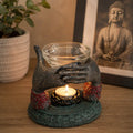 Antique Buddha - Offering Hands Oil Burner showing a decorative resin Buddha hands sculpture holding a glass bowl with a lit tealight candle beneath, placed on a wooden surface with a Buddha print and plant in the background.