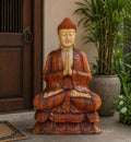 Hand Carved Buddha Statue - 80cm Welcome sitting peacefully with hands in prayer pose, beautifully crafted from rich wood, placed near a wooden door and green plants.
