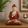 Hand Carved Buddha Statue - 30cm Teaching Transmission seated in meditation with detailed red and beige robe, placed on a wooden surface with a plant, candle, and framed artwork in the background.