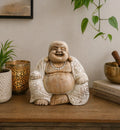 Happy Buddha - Whitewash 30cm sitting on a wooden surface surrounded by a plant, lit candle in a decorative holder, and stacked books, showcasing a hand carved Buddha statue with a serene smile.