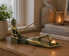 Antique Buddha - Hand Incense Burner displayed on a wooden surface with a gold-toned Buddha figure holding a lit candle, burning incense stick releasing smoke, surrounded by cozy home decor.