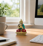 Brass Buddha Figure - Blessing - 7.5cm sitting on a wooden desk with a turquoise and red inlay design, surrounded by a plant, coffee mug, keyboard, and computer monitor in a bright workspace.