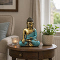 Teal & Gold Buddha - Large resin Buddha statue sitting in meditation pose on a wooden side table, styled with a potted plant and lit candle in a cozy living room setting.