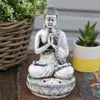 White resin Buddha Candle Holder - White - Medium with a detailed seated Buddha figure meditating, holding hands in prayer above a small lit tea light candle on a rustic wooden surface.