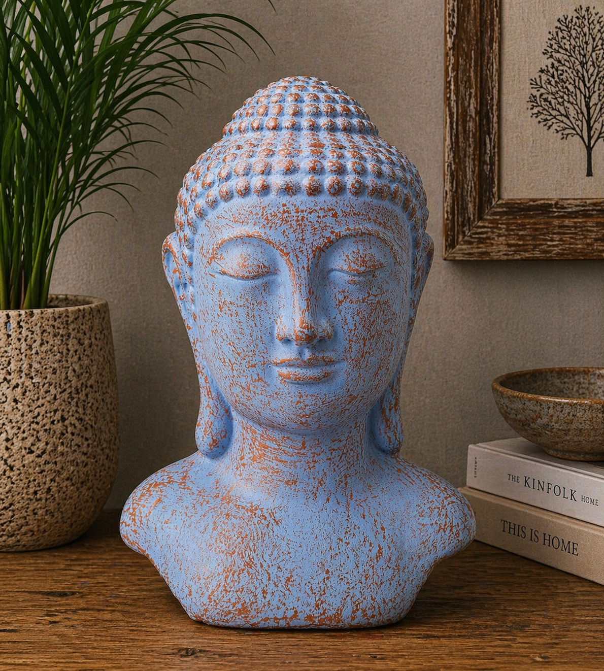 A blue and rust-colored Buddha head statue with closed eyes, placed on a wooden surface next to a plant in a textured pot and stacked books under a framed wall art.
