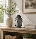 A serene Home Decoration Buddha Head - 25cm - Silver Shine Finish displayed on a wooden table beside a ceramic vase with greenery and stacked books in a calm, natural light setting.
