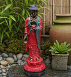 Antique Buddha - Standing statue in vibrant red and teal hues, placed outdoors on a stone surface surrounded by green plants and natural decor.