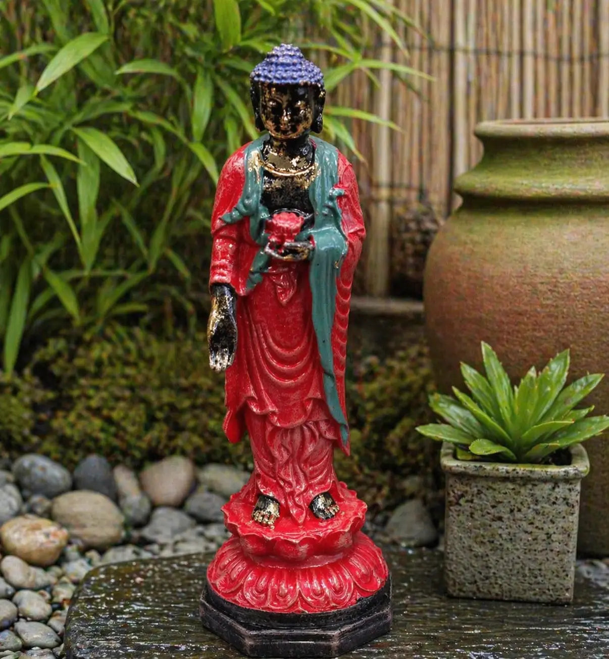 Antique Buddha - Standing statue in vibrant red and teal colors with a blue hair design, displayed outdoors among green plants and decorative pots, creating a serene garden atmosphere.